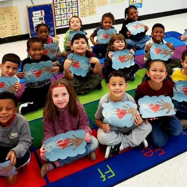 Photo of students sitting on a carpet holding their art work.